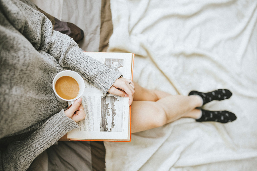 白いシーツのベッドの上で、本を開きコーヒーカップを手にしているが、飲むでも読むでもない様子の女性の写真