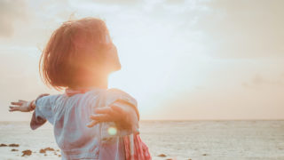 太陽の明るい光が溢れる海辺で両手を広げて大空を見つめている女性の後ろ姿の写真