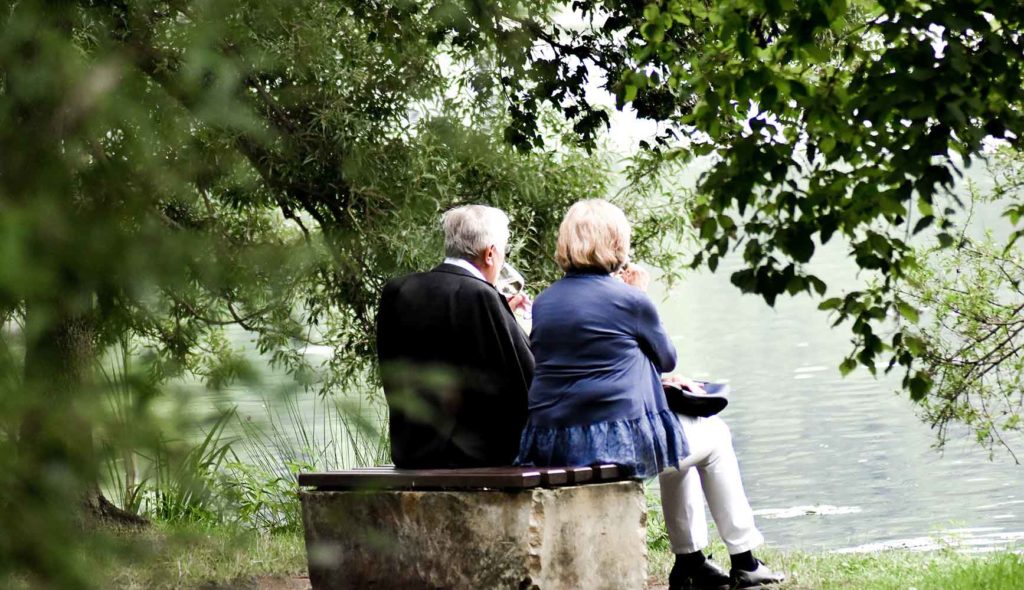 グリーンが綺麗な湖のほとりで二人並んで座っている老夫婦の後ろ姿の写真