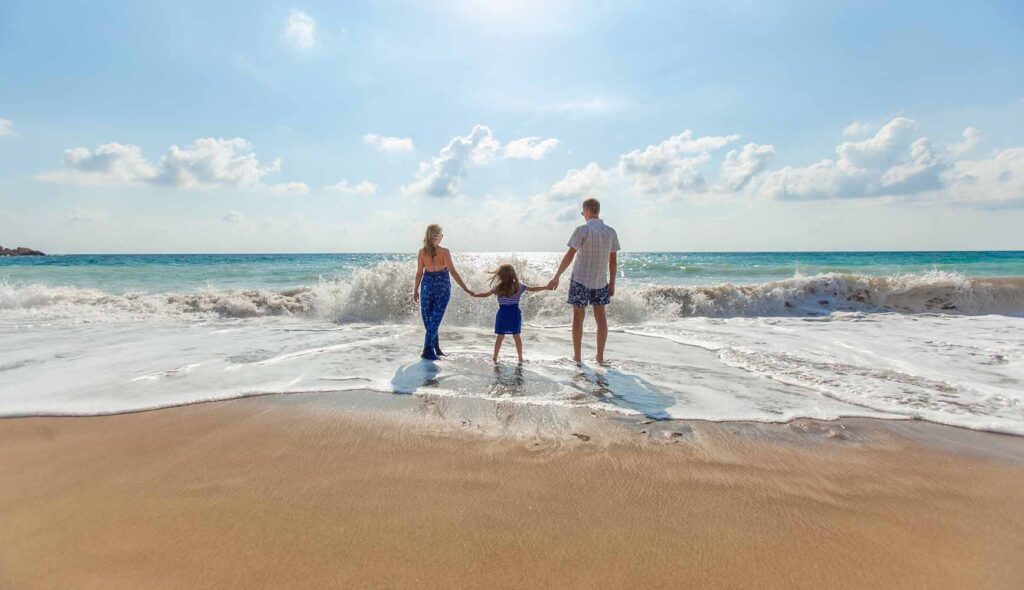 晴天の海を見つめ、５歳くらいの女の子を真ん中に三人で手を繋ぐ、ブルーのサマードレスがおしゃれな妻のいる家族の後ろ姿の写真