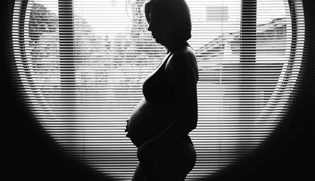 窓際で俯き加減でお腹に手を添える妊婦のモノクロ写真